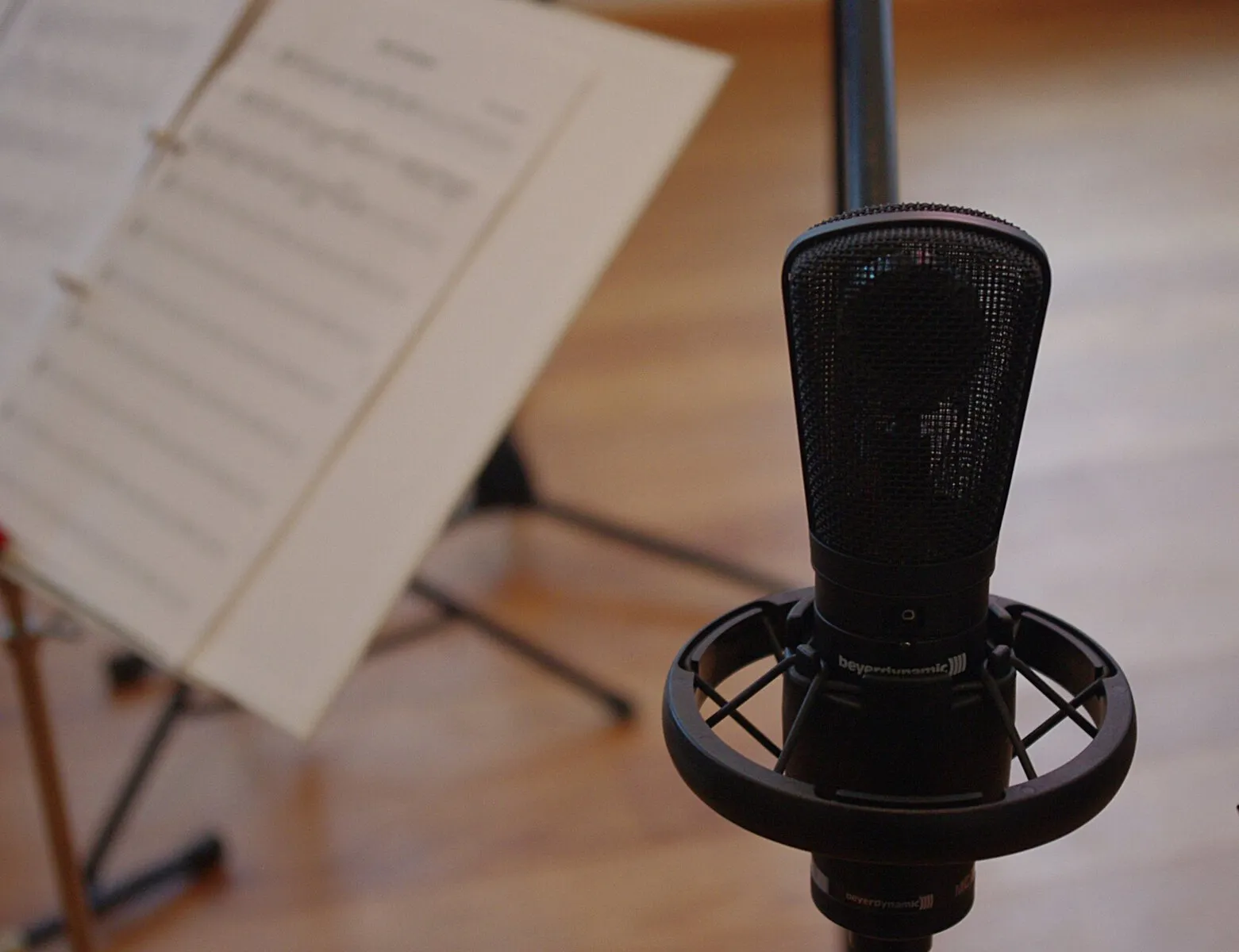 A studio microphone on a stand beside a music stand holding open sheet music, in a lesson room with a warm wood floor.